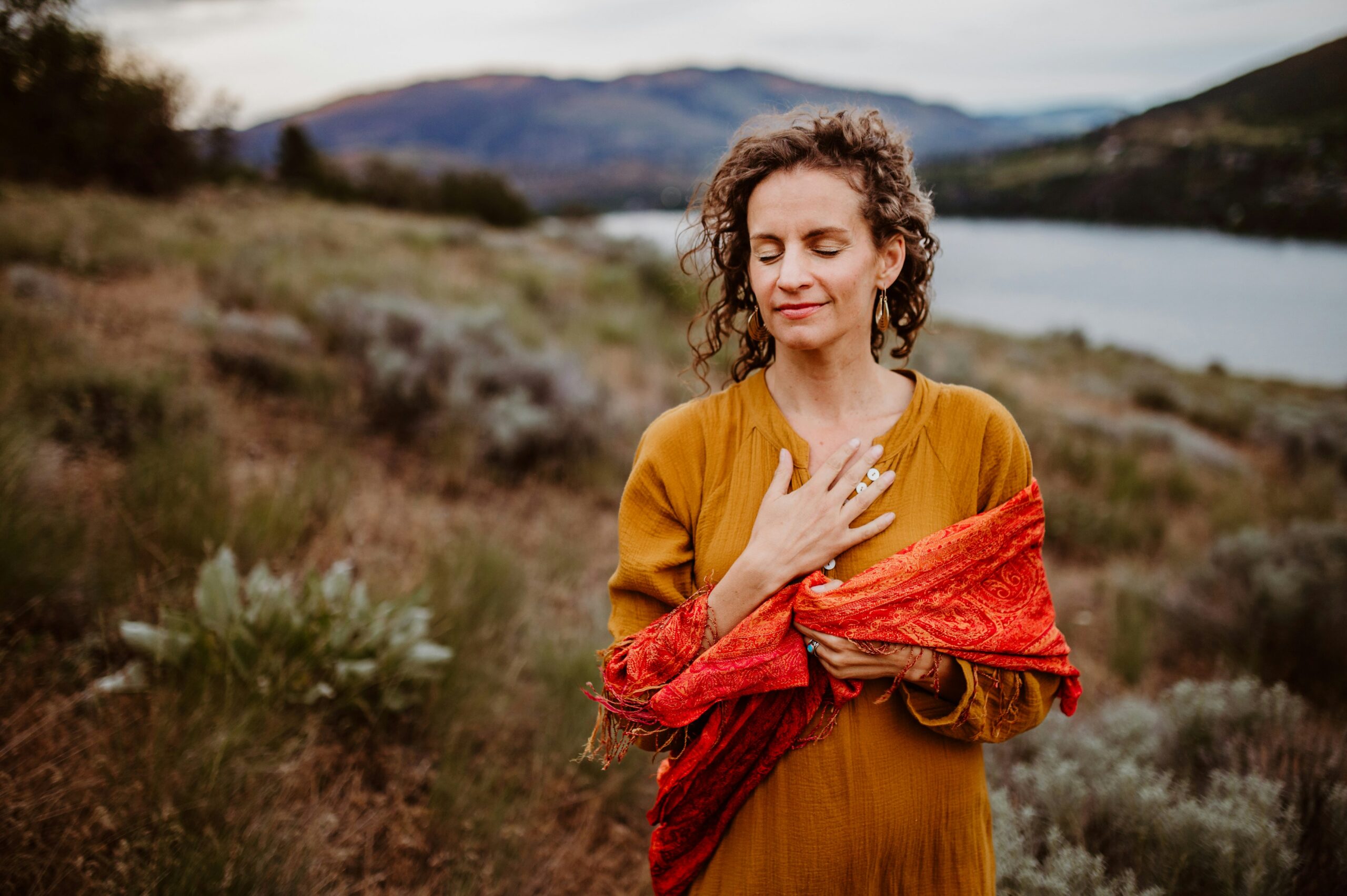 amie-roussel-BCxM9FStGrs-unsplash Woman standing on the grassy shores of Kalamalka Lake with her hand over her heart and eyes closed in thoughtful meditation and connection with nature - Rachel Wyatt Creative by Amie Roussel